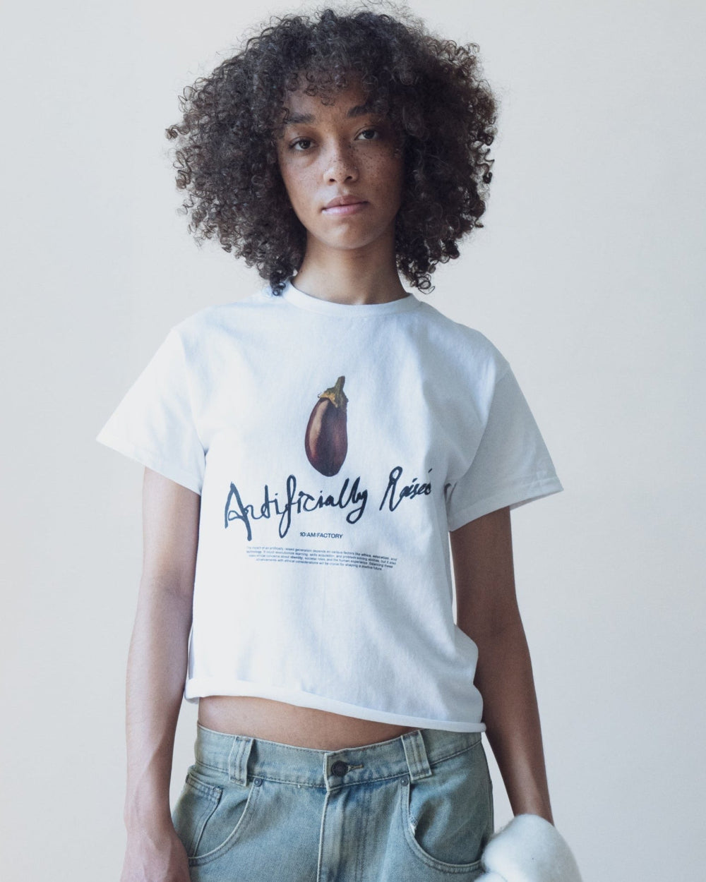 Unisex white 'Artificially Raised' t-shirt by 10:AM Factory, featuring a bold big eggplant graphic print and 10:AM logo, crafted from high-quality, soft cotton for a clean, modern look.